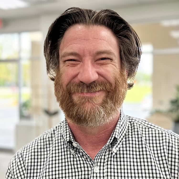 Bearded man smiles at the camera, standing indoors near large windows in a brightly lit room; wearing a black-and-white checkered shirt with medium-length hair swept back.