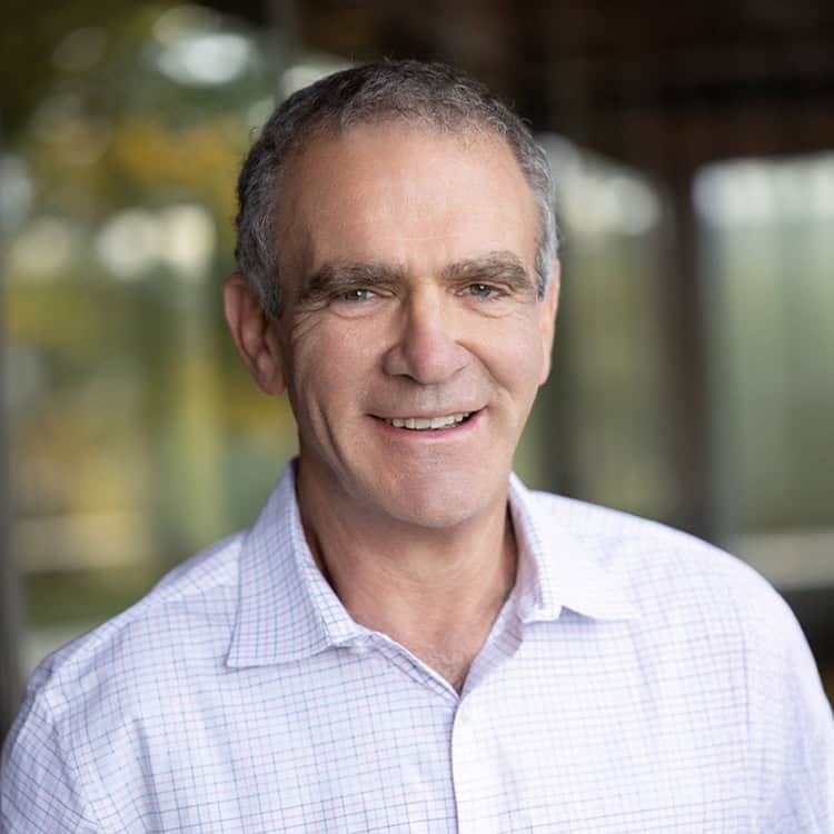 Middle-aged man smiles at camera, wearing a light checked dress shirt; standing indoors near large blurred windows with soft natural light and out-of-focus greenery behind him.
