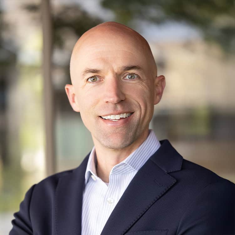 A bald man smiles at the camera, wearing a navy blazer and striped dress shirt, standing before blurred glass and greenery, arms folded.