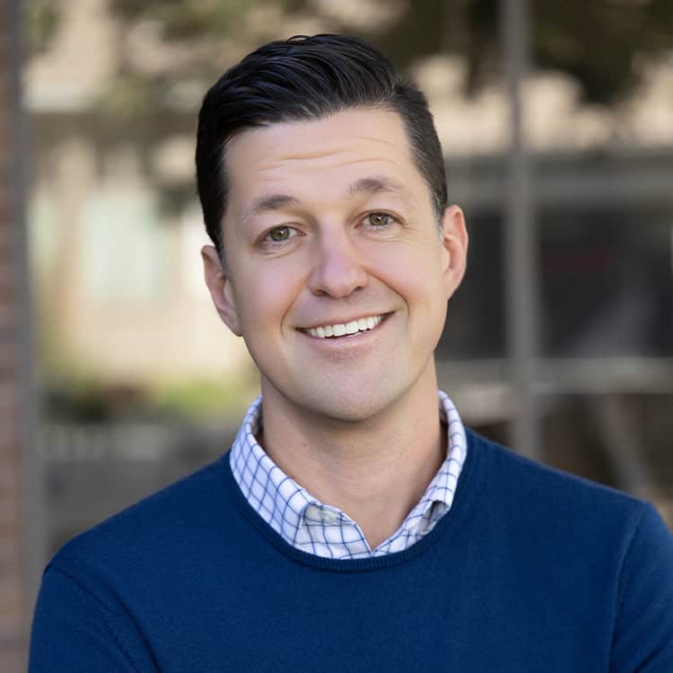 Adult man smiling directly at camera, wearing a blue sweater over a checked shirt, standing outdoors in front of a blurred urban background with windows and brick.