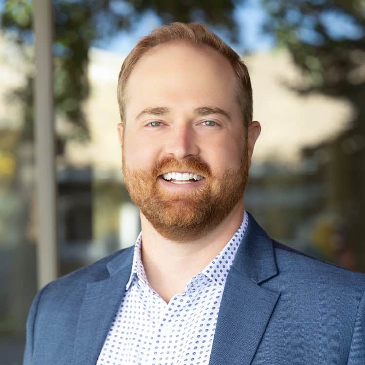 Smiling bearded man looks toward the camera, wearing a blue blazer and white patterned shirt; standing outdoors near reflective glass with blurred trees and a building in the background.