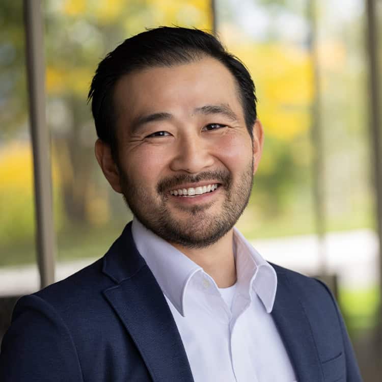 Person with short dark hair and trimmed beard smiling at camera, wearing a navy blazer and white collared shirt, standing in front of blurred glass with autumnal outdoor background.