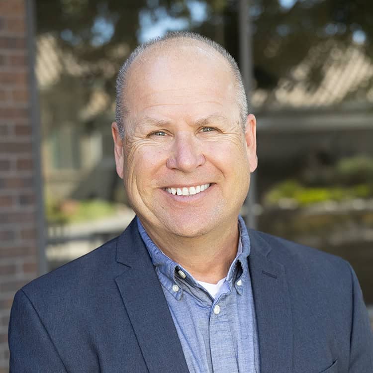 Smiling middle-aged bald man looks at camera, wearing a navy blazer and light blue button-down shirt, standing outdoors before a blurred building facade with trees and reflected windows.