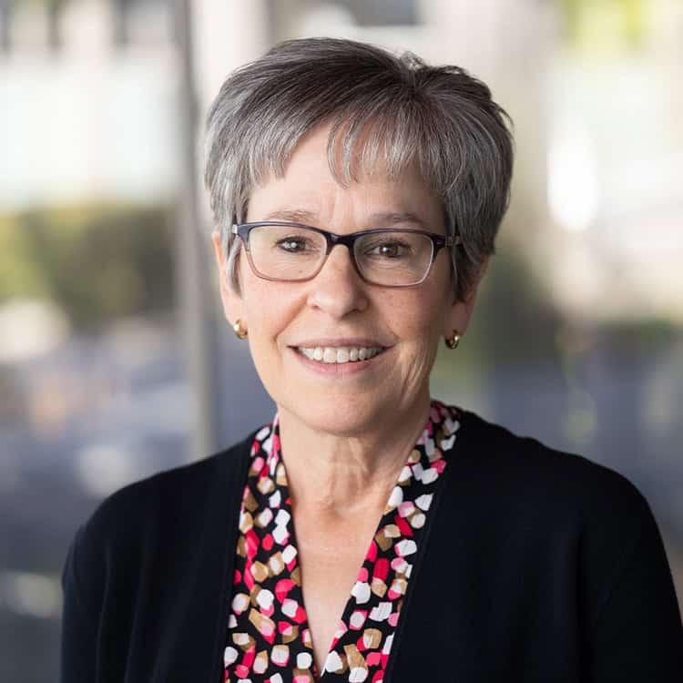 A gray-haired woman wearing glasses smiles at the camera; she wears a black cardigan over a colorful patterned blouse, standing before a softly blurred indoor windowed background.