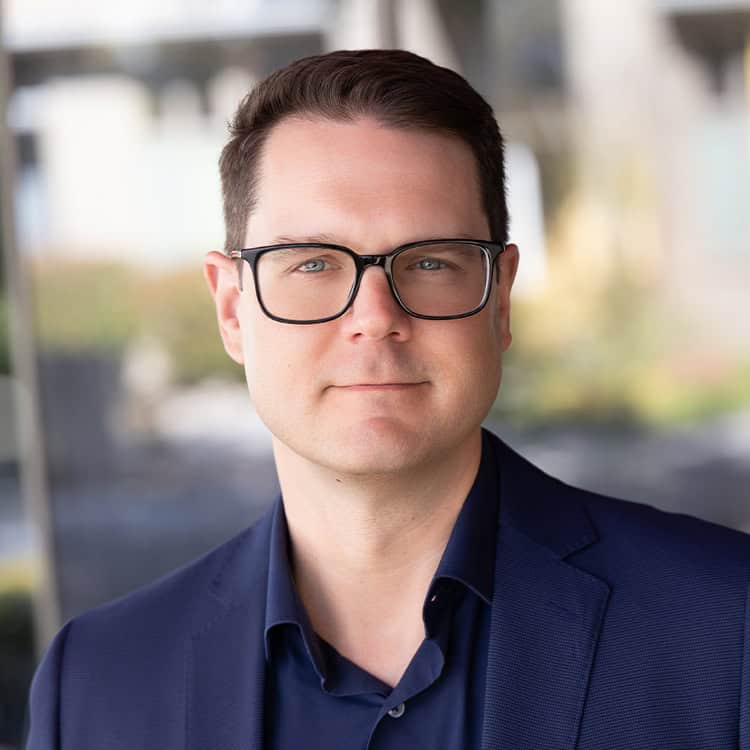 Adult man wearing black-framed glasses and a navy blazer looks directly at the camera with a neutral expression; shallow depth-of-field shows a softly blurred outdoor, urban background.