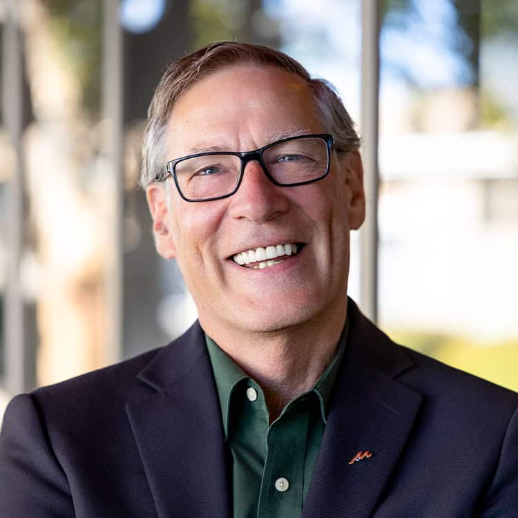 Smiling older man with glasses and short gray hair, wearing a dark blazer and green shirt, stands indoors near a window with a bright, blurred outdoor background.