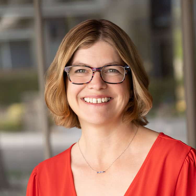 Woman with short light-brown hair and patterned glasses smiles at the camera, wearing a red blouse and delicate necklace against a softly blurred windowed background.