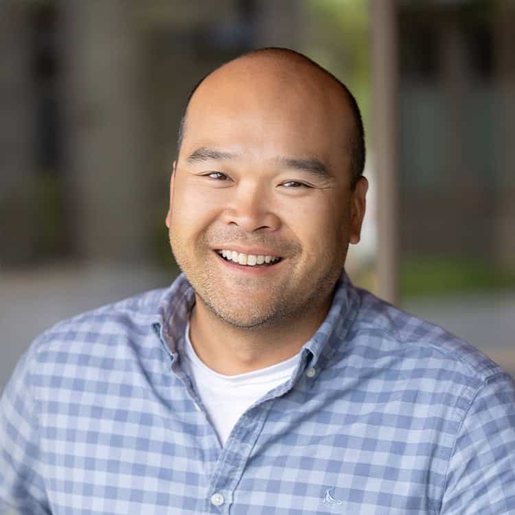 Person smiling directly at camera, wearing a blue checkered button-up shirt and white undershirt; sitting outdoors with blurred columns and greenery in a soft-focus background.