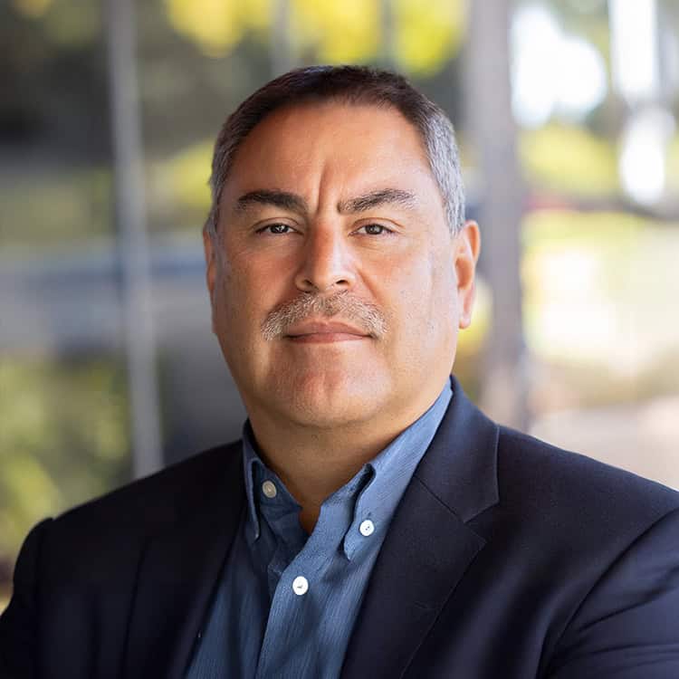 Middle-aged man with salt-and-pepper hair and mustache, looking directly at the camera, wearing a navy blazer and blue button-up, standing before a blurred outdoor glass-and-greenery background.