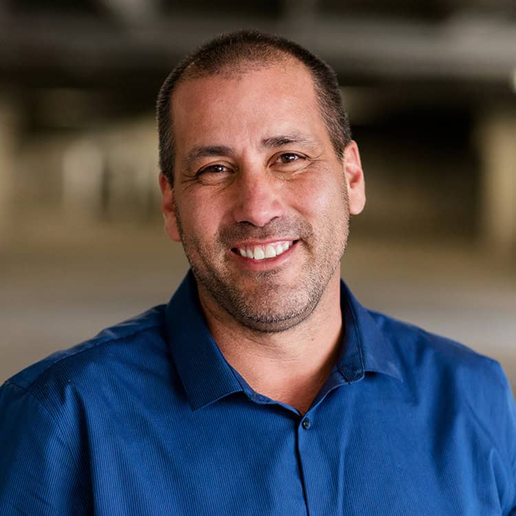 Adult man smiles at the camera, wearing a blue collared shirt; short hair and light stubble. Context: softly blurred, neutral-toned interior background.