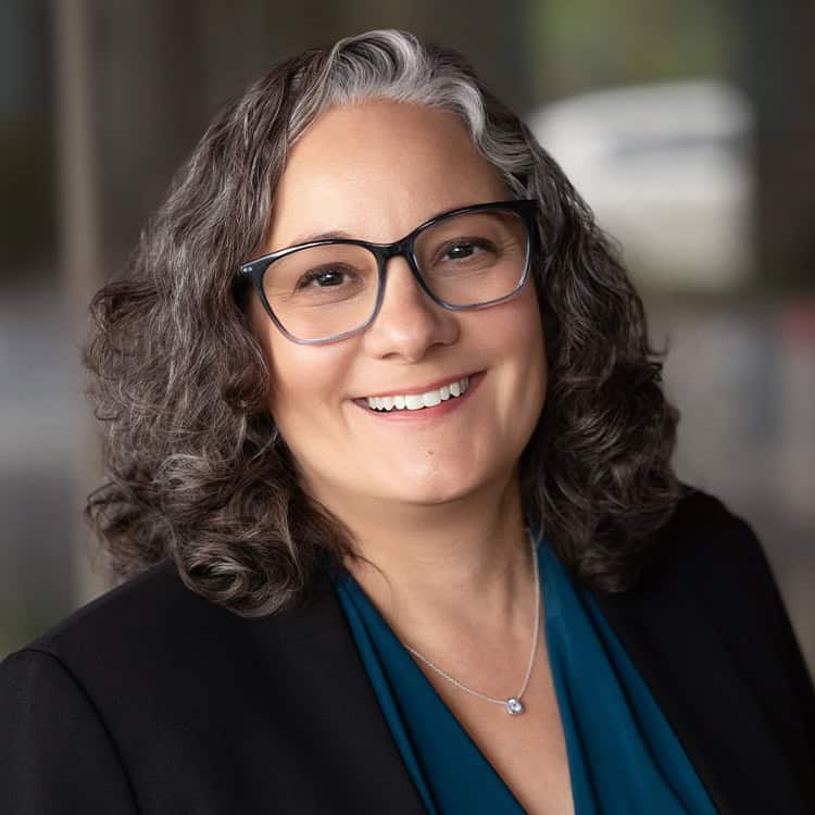 A smiling woman with gray-streaked curly hair and black glasses looks at the camera, wearing a teal blouse, black blazer and a small pendant necklace; soft, blurred indoor background.
