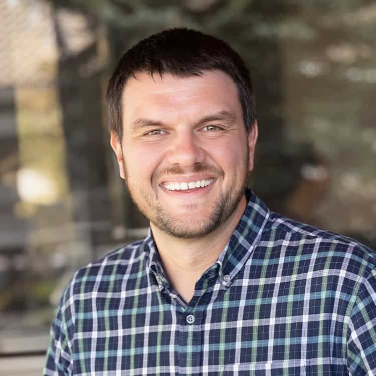 A smiling adult man with short dark hair and light stubble looks at the camera, wearing a blue-green plaid button-up, standing before a blurred glass and tree background.