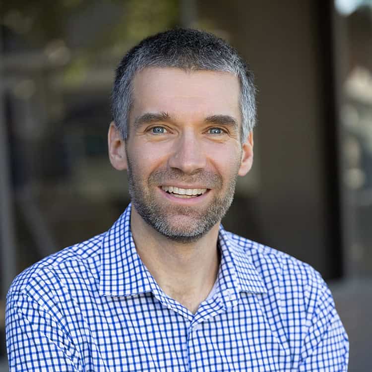 Smiling man with short salt-and-pepper hair and light beard, wearing a blue checkered shirt, looking at the camera while standing before a softly blurred outdoor/architectural background.