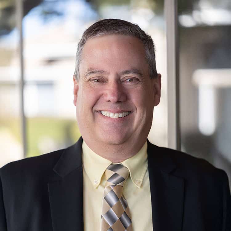 Middle-aged man smiling warmly, wearing a dark suit, pale yellow shirt and patterned tie, standing indoors beside a glass window with a softly blurred outdoor background.