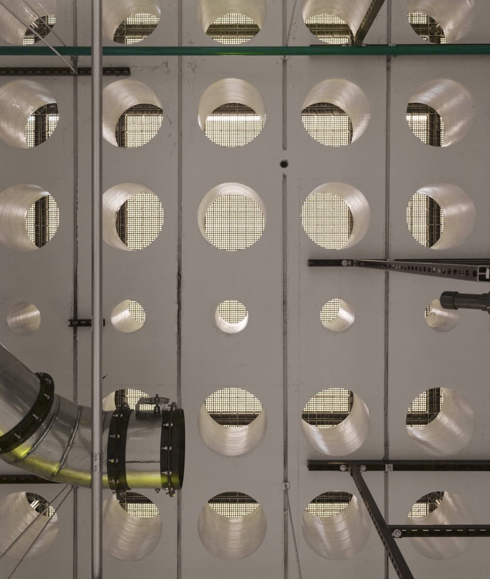Perforated ceiling panel with repeated circular cutouts showing backlit mesh; a metal duct and vertical support rods run across the openings, set in an industrial mechanical room ceiling.