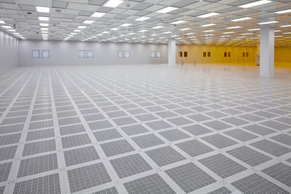 Raised perforated floor tiles form a grid, reflecting ceiling lights across an empty, spacious cleanroom with white walls, columns, small windows and a warm yellow-lit area to the right.