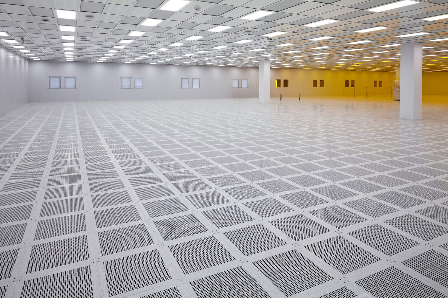 Raised perforated floor tiles form a grid, reflecting ceiling lights across an empty, spacious cleanroom with white walls, columns, small windows and a warm yellow-lit area to the right.