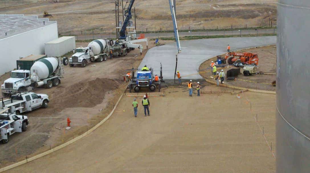 Construction crew pouring and smoothing a curved concrete slab at an open industrial site with cement mixers, pump truck, utility vehicles, and a large storage tank nearby.