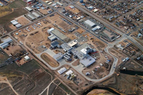 A large industrial complex sprawls with interconnected white-gray buildings, storage tanks, loading bays and parked trucks amid curved access roads, bordered by residential streets and open fields.