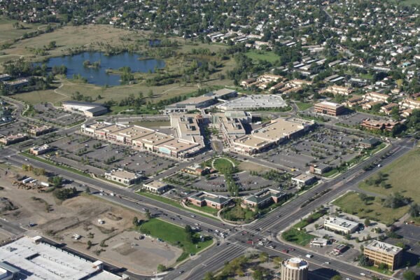 A large suburban shopping mall sits amid expansive parking lots with scattered cars, intersected by busy multi-lane roads, bordered by residential neighborhoods and a small pond in the background.