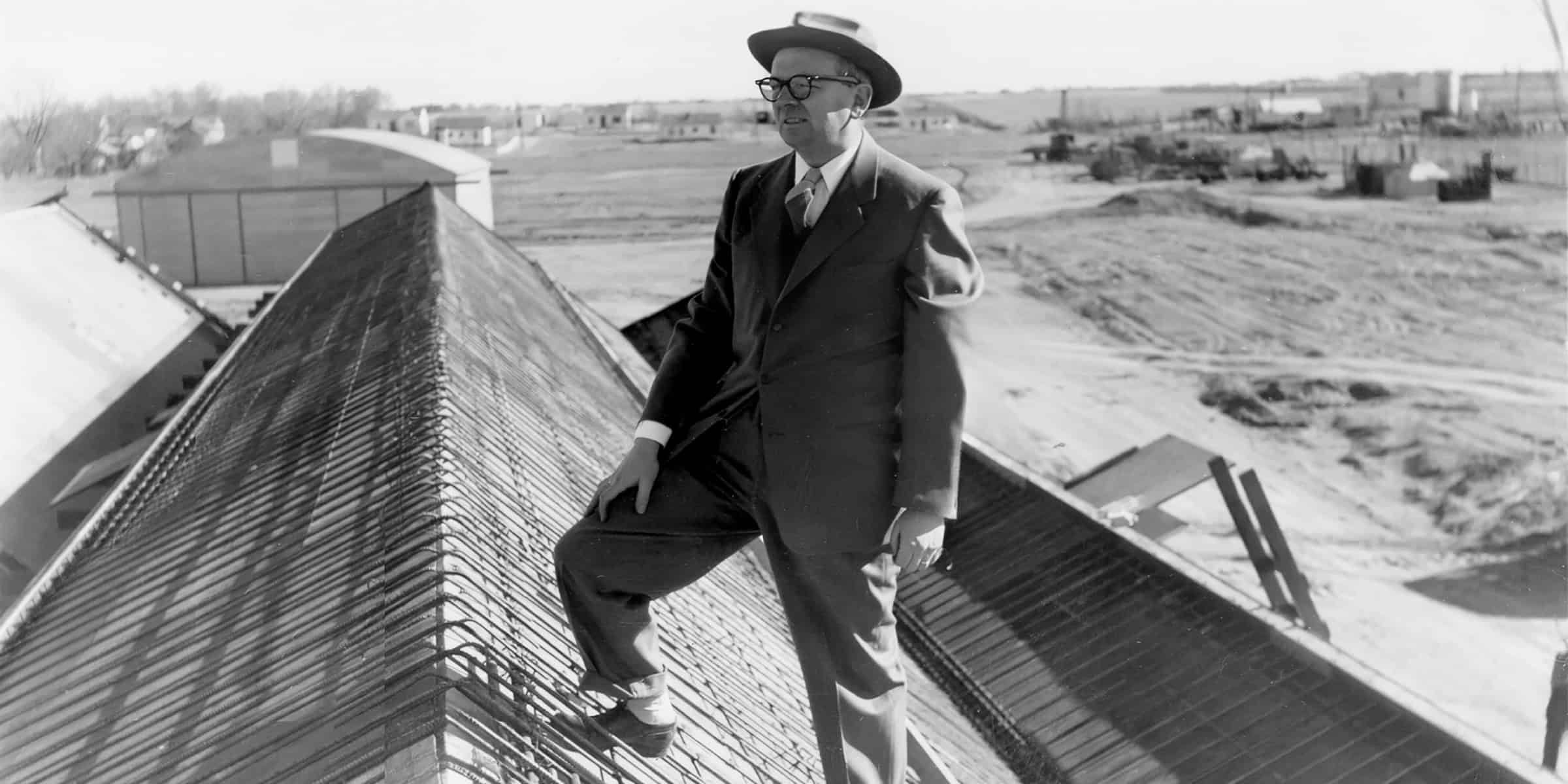 Well-dressed man in suit and hat stands with one foot on a metal roof ridge, gazing across a flat, sparsely built landscape of hangars, small buildings and dirt roads.