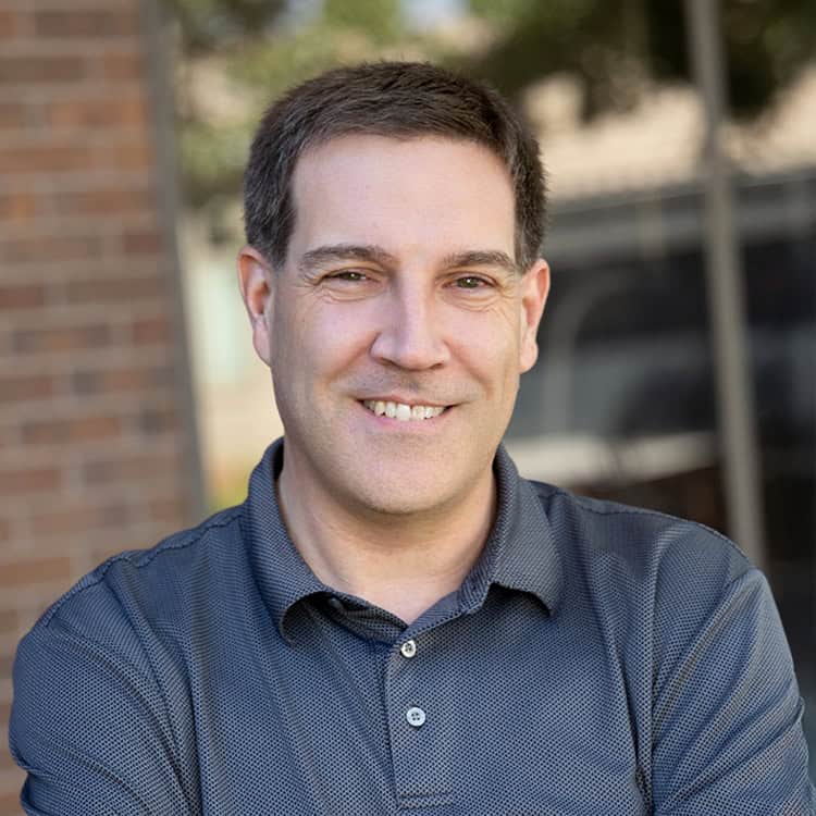 Middle-aged man smiling with arms crossed, standing outdoors by a brick wall and glass window; wearing a dark short-sleeve polo, short brown hair, light stubble, relaxed expression in soft daylight.