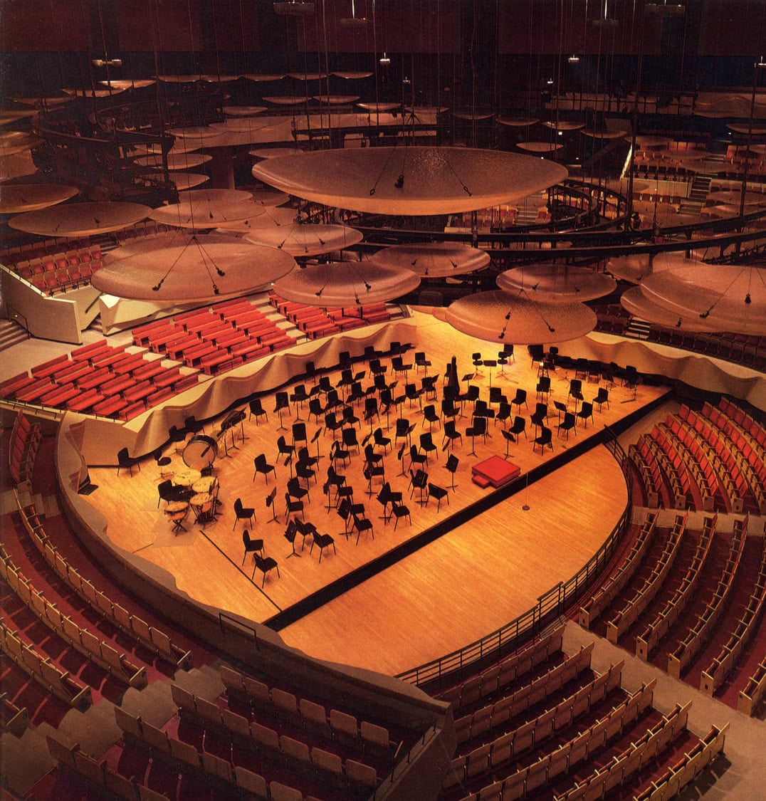 Orchestra stage with empty chairs, music stands and percussion arranged for performance; large suspended circular acoustic panels hang above; surrounded by curved, tiered red-and-beige seating in a modern concert hall.