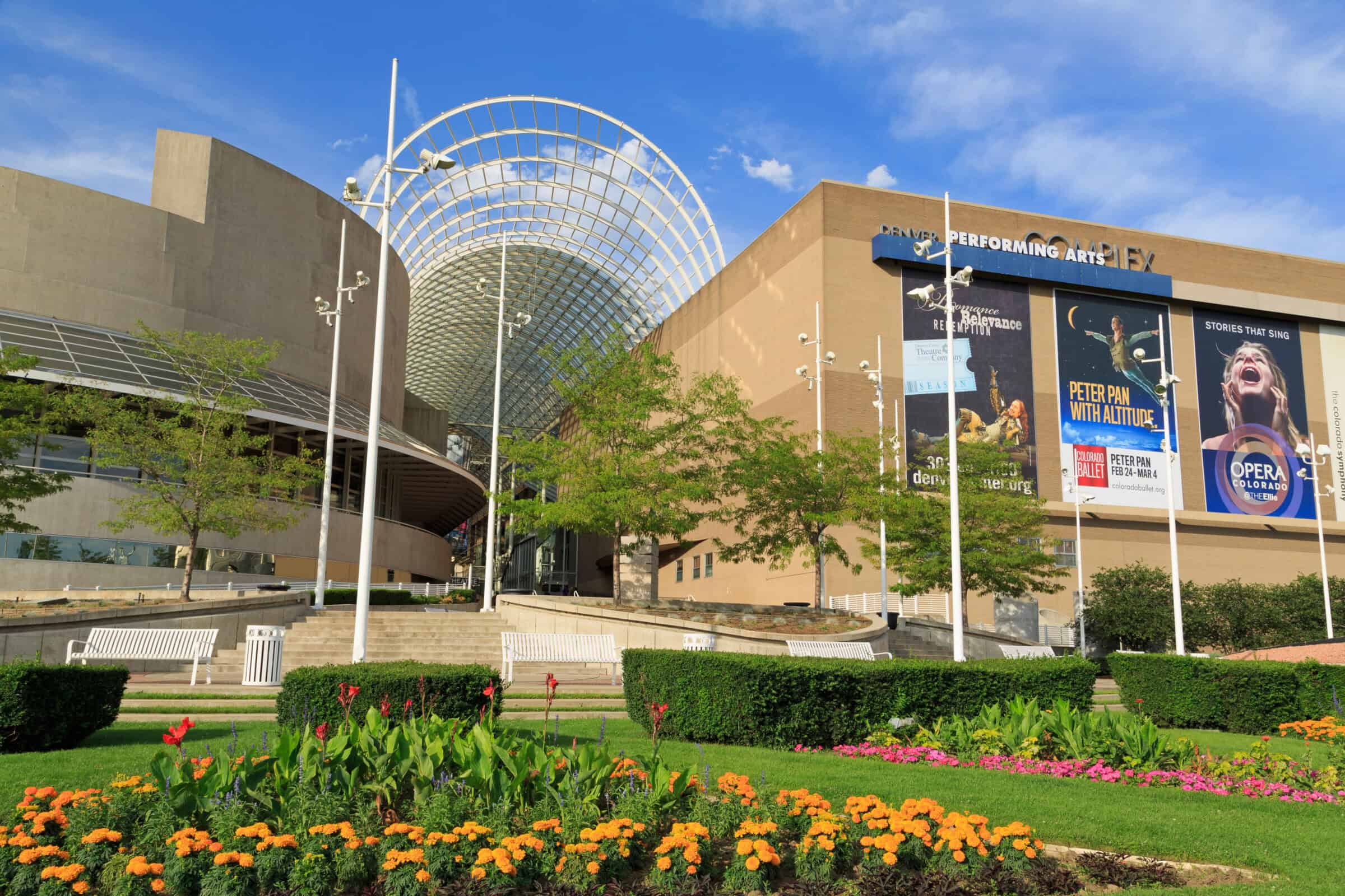 Modern performing-arts complex (object) displays a large arched glass canopy and advertising posters (action) amid manicured flowerbeds, benches, and clear blue sky (context).

Text visible: "DENVER PERFORMING ARTS"; "PETER PAN WITH ALTITUDE"; "COLORADO BALLET"; "PETER PAN FEB 24 - MAR 4"; "coloradoballet.org"; "STORIES THAT SING"; "OPERA COLORADO"; "the colorado symphony"