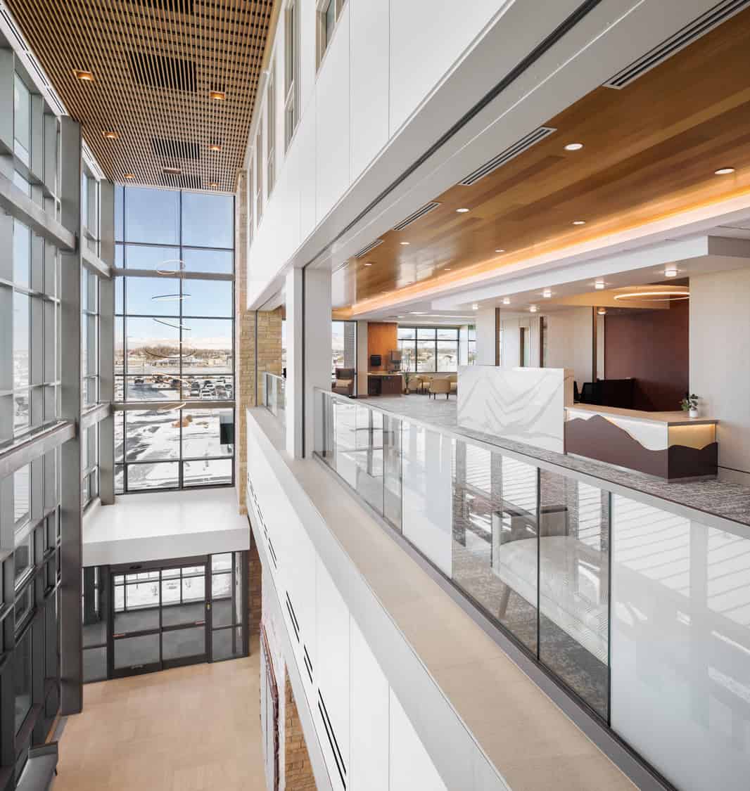 Glass-railed mezzanine overlooks a modern reception area; warm wood ceilings and circular pendant lights illuminate seating and a marble desk, framed by floor-to-ceiling windows showing a snowy parking lot outside.