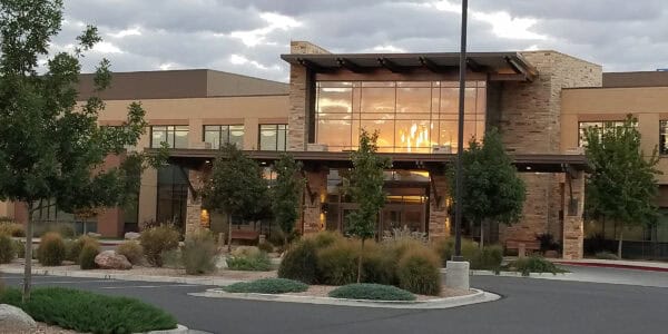 A modern stone-and-glass building reflects a golden sunset through its large windows, framed by trees and ornamental grasses in a landscaped parking lot under a cloudy sky.