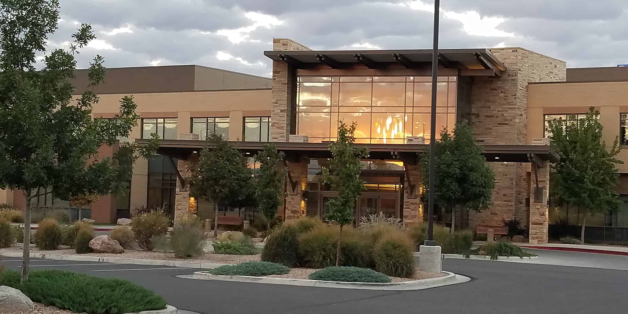 A modern stone-and-glass building reflects a golden sunset through its large windows, framed by trees and ornamental grasses in a landscaped parking lot under a cloudy sky.