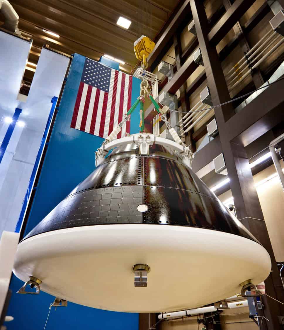Space capsule suspended by straps and a yellow hoist, being lowered inside a tall industrial assembly bay with an American flag on a blue wall and exposed structural beams.