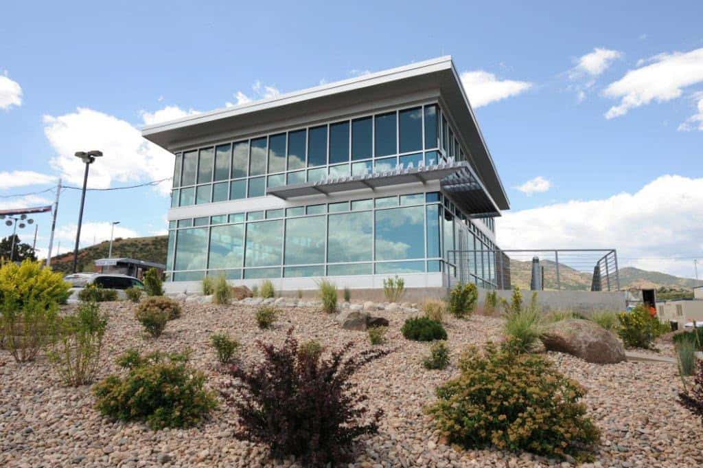 A two-story modern glass building sits atop a rocky, drought-tolerant landscaped slope, overlooking parked cars and distant hills under a partly cloudy blue sky.