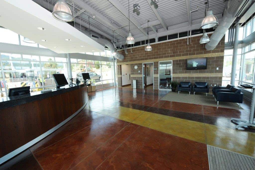 Curved reception desk holds computer monitors in a spacious modern lobby with polished multicolored concrete floor, leather seating, large windows and exposed industrial ceiling lights.