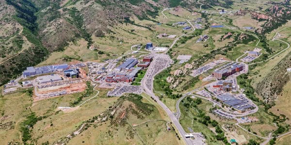 Sprawling campus of buildings and parking, aligned along a central road; parked cars fill lots while winding roads cross the site, surrounded by rolling green hills and rocky outcrops.