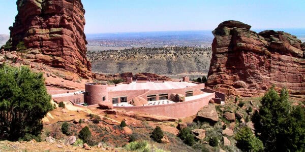 A low, rectangular brick building sits nestled between two towering red sandstone cliffs, surrounded by scrubby vegetation and rocky outcrops, overlooking a distant valley and scattered suburban development.