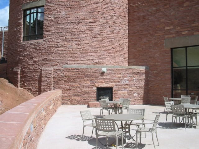 Metal patio tables and chairs sit arranged on a sunlit concrete patio beside a curved red sandstone building with tall windows and a low stone wall.
