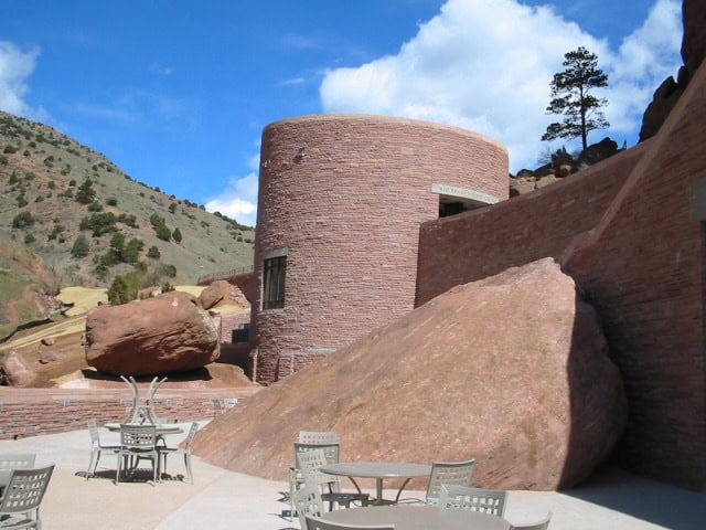 Cylindrical red‑stone building nestled against massive reddish boulders, with metal patio tables and chairs arranged on a paved terrace, set in a rocky hillside under a bright blue sky.