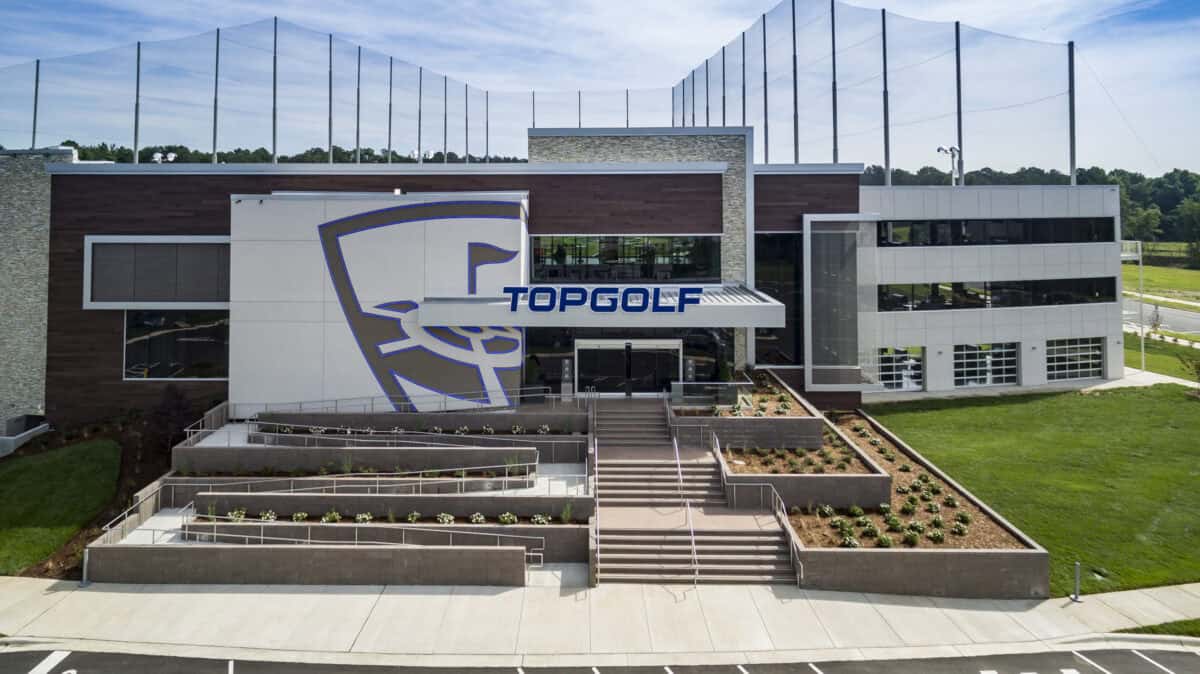 Topgolf building displays large logo and sign reading "TOPGOLF" above the main entrance, with terraced planters and stairways leading to glass doors, flanked by driving-range netting and grassy grounds.