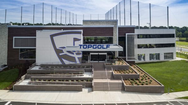 Topgolf building displays large logo and sign reading "TOPGOLF" above the main entrance, with terraced planters and stairways leading to glass doors, flanked by driving-range netting and grassy grounds.