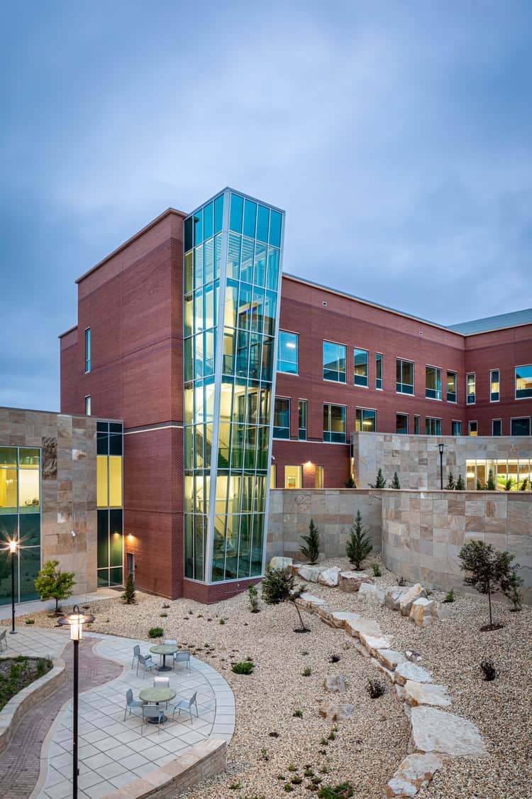 Glass stair tower of a modern red-brick building glows with interior lights, revealing a spiral staircase; empty patio tables sit in a rock‑landscaped courtyard under a cloudy evening sky.