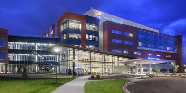 Children's hospital building — illuminated at dusk, glass-fronted main entrance with canopy, landscaped lawn and curved driveway under a deep blue evening sky. Text: "Children's Hospital Colorado Colorado Springs" and "CHILDRENS MAIN ENTRANCE".
