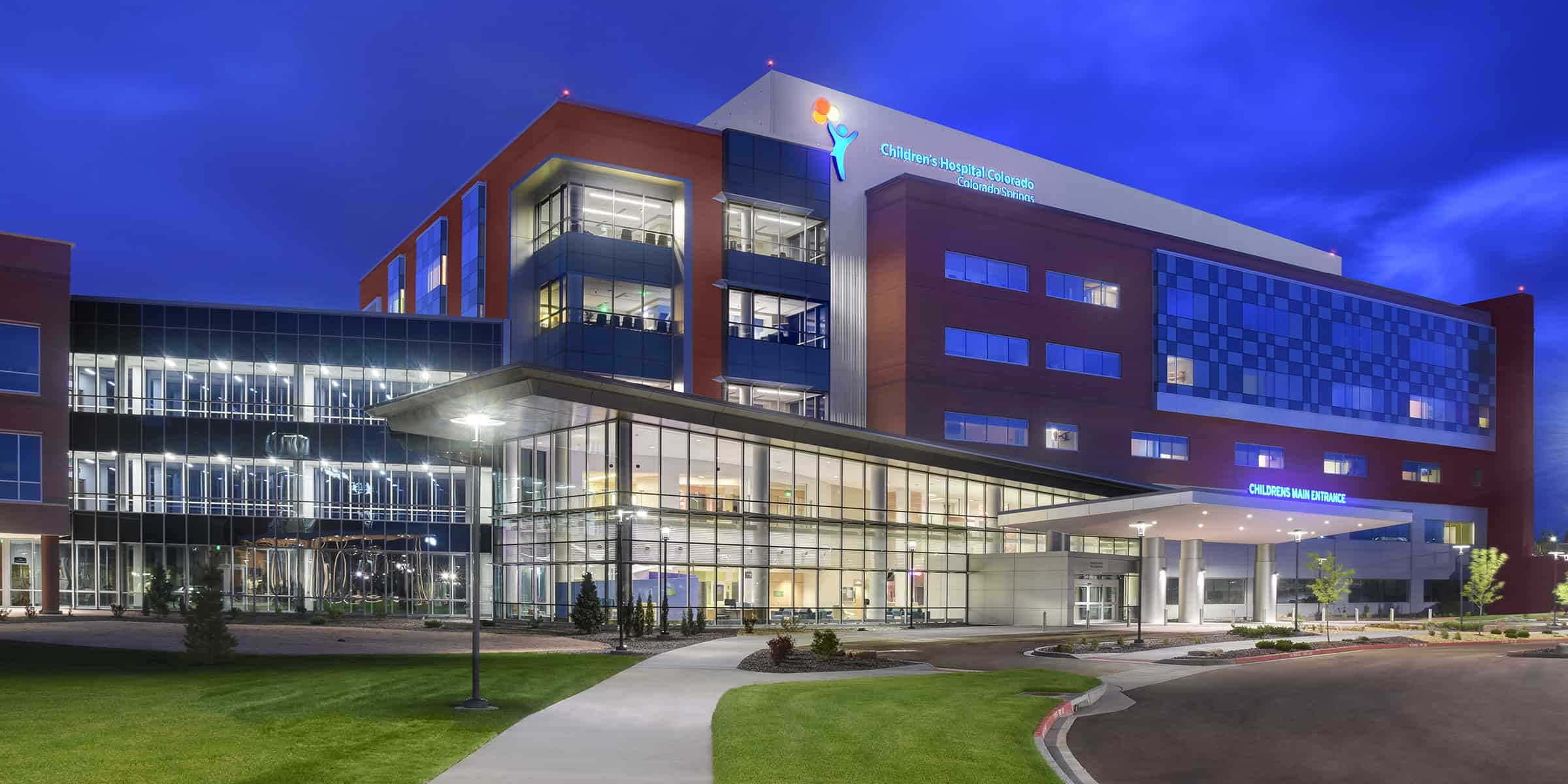 Children's hospital building — illuminated at dusk, glass-fronted main entrance with canopy, landscaped lawn and curved driveway under a deep blue evening sky. Text: "Children's Hospital Colorado Colorado Springs" and "CHILDRENS MAIN ENTRANCE".