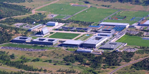 Sprawling institutional campus sits around a large central lawn, with office buildings, parking lots, tennis courts and baseball fields, on a grassy plateau surrounded by roads and pine-covered hills.