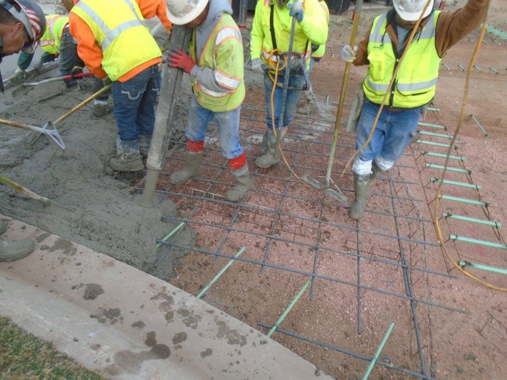 A crew of construction workers pouring and leveling wet concrete onto a steel rebar grid with a chute and rakes, wearing high-visibility vests and hard hats beside a curb.