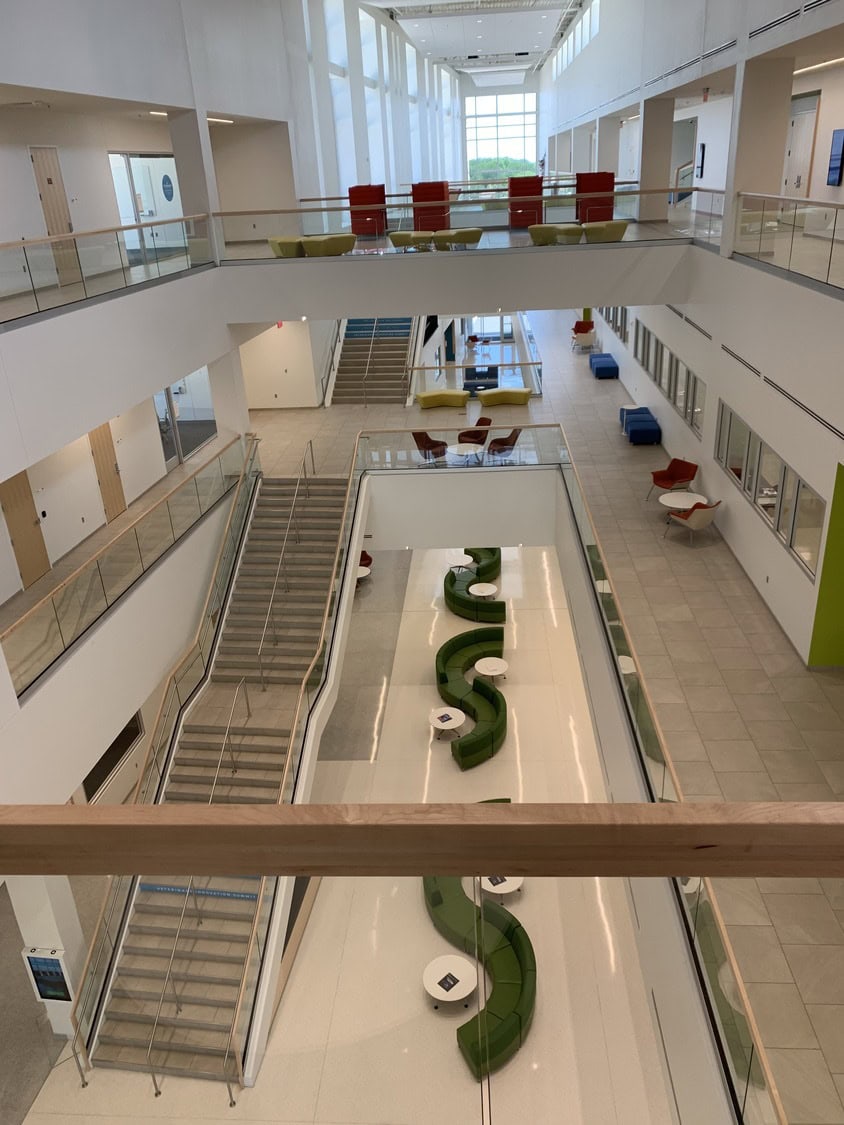 Atrium hosts a sinuous row of green sofas down the central corridor; staircases connect three levels; red and yellow seating punctuates glass-railed balconies; tall windows flood the white interior with daylight.