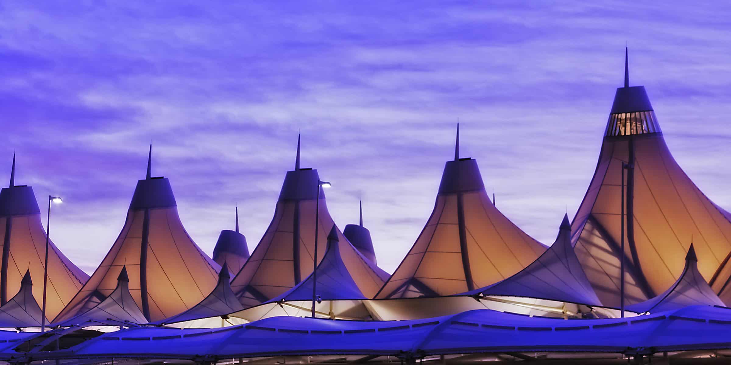 A series of fabric-cone roof peaks glow warm orange from internal lighting, rising like tents against a soft purple-blue twilight sky above a modern terminal-like structure.