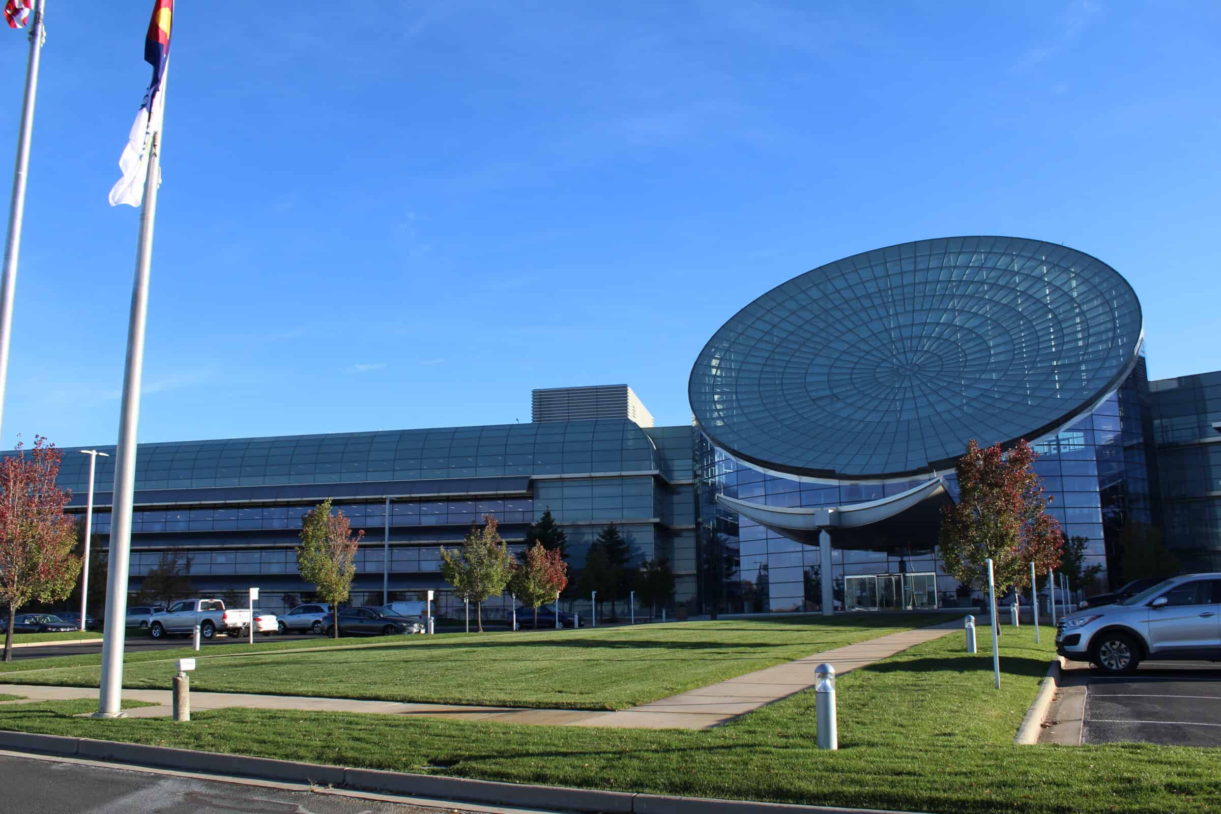 Glass office building with a large circular glass canopy tilting over its main entrance, standing amid a manicured lawn, parked cars, flagpoles, and a clear blue sky.