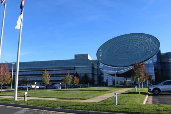 Glass office building with a large circular glass canopy tilting over its main entrance, standing amid a manicured lawn, parked cars, flagpoles, and a clear blue sky.