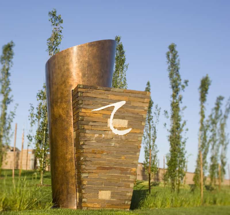 Stone-and-copper monument displays a white stylized "Z" while standing on a manicured lawn, backed by tall slender trees and a clear blue sky.